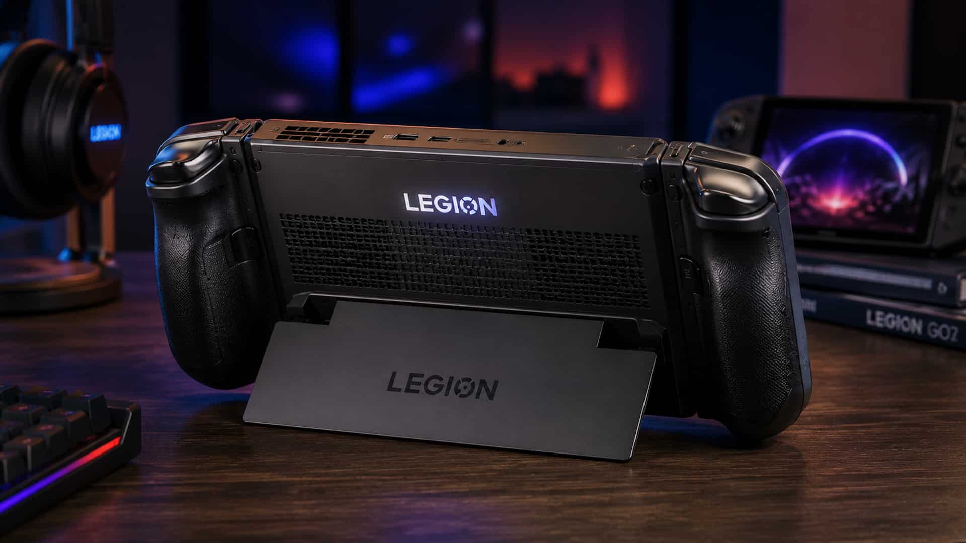 Das Bild zeigt die Rückseite eines Gaming-Handhelds im Stil der Lenovo Legion Go 2. Das Gerät steht auf einem ausgeklappten Standfuß auf einem Schreibtisch. Sichtbar sind die Lüftungsöffnungen, die Griffe auf der Rückseite sowie ein beleuchtetes Legion-Logo. Im Hintergrund befinden sich unscharf weitere Gaming-Geräte und Zubehör.